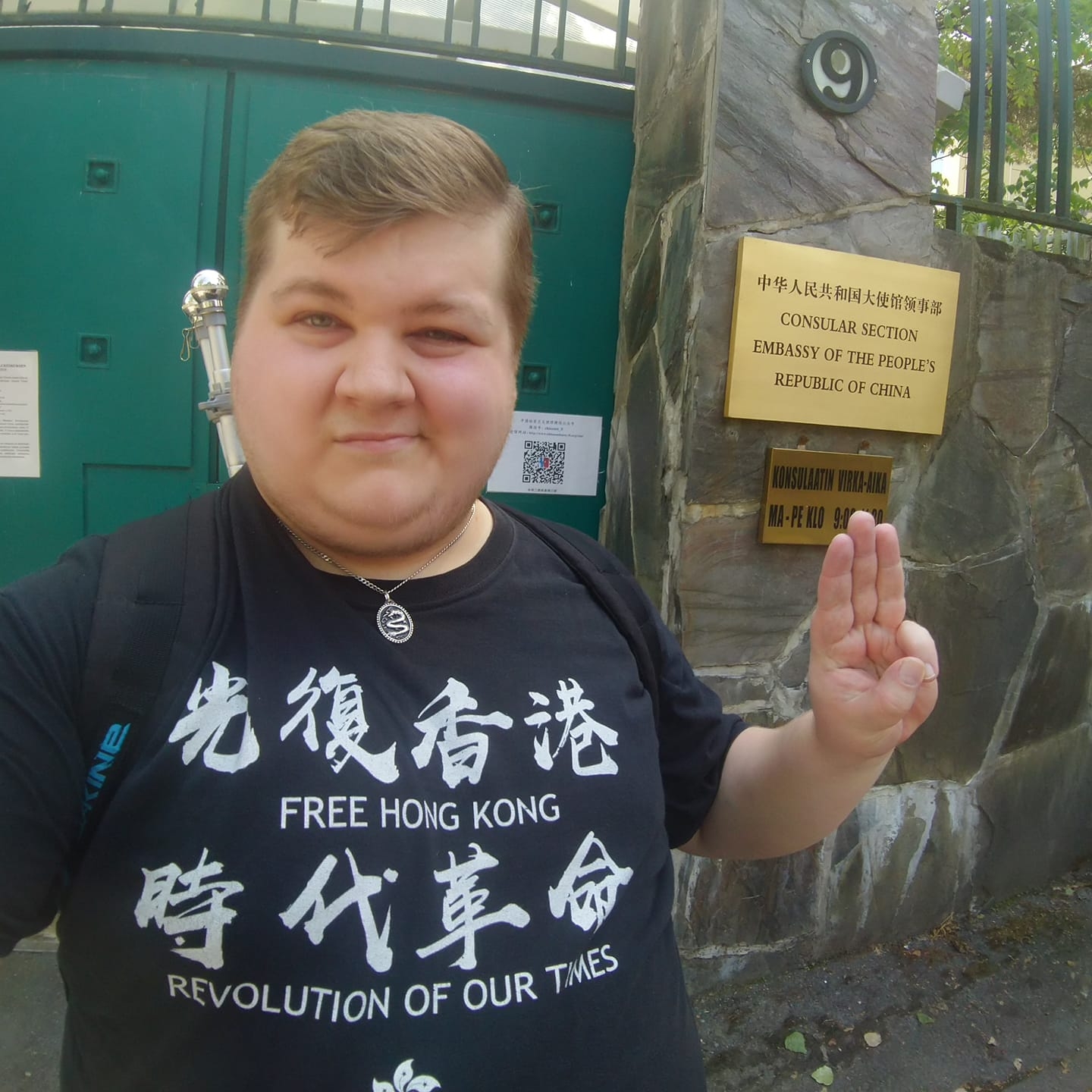 Eero Kivistö seisoo rakennuksen koristeellisella portilla ja tervehtii kolme sormea pystyssä. Paidassa lukee: Free Hongkong!