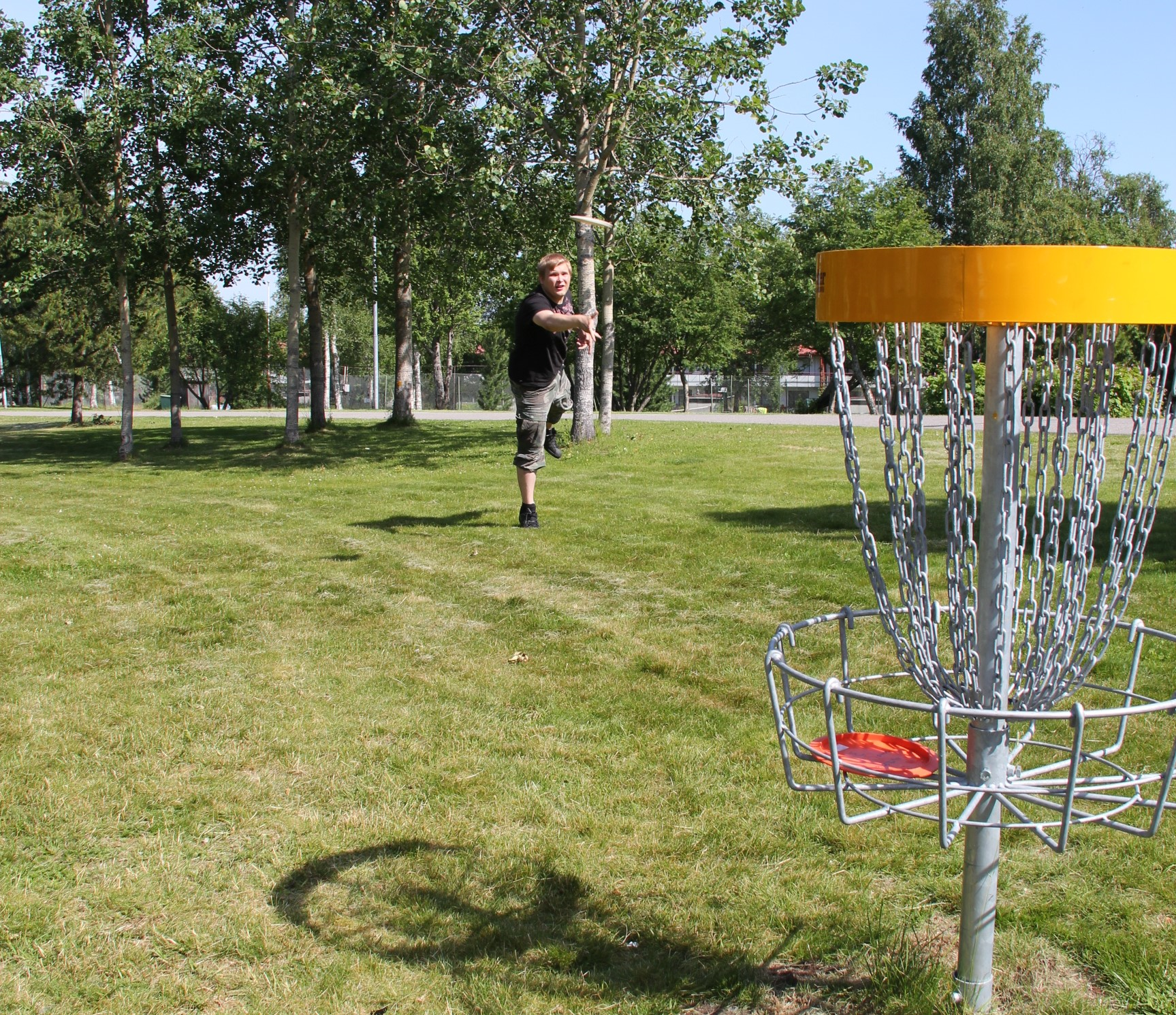 Elias Helminen heittää frisbeetä koriin aurinkoisella kentällä