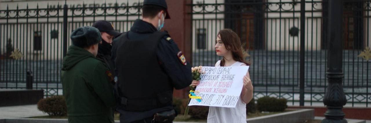 Valkoisiin pukeutunut nainen seisoo kadulla pitäen kädessään venäjänkielistä tekstiä sisältävää kylttiä.