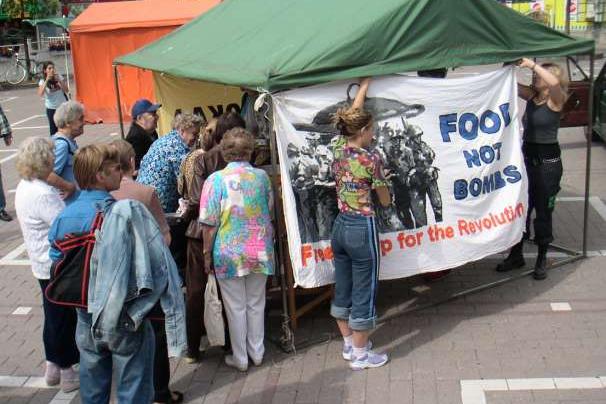 Kaksi vapaaehtoista kiinnittää Food Not Bombs -julistetta teltan kylkeen.
