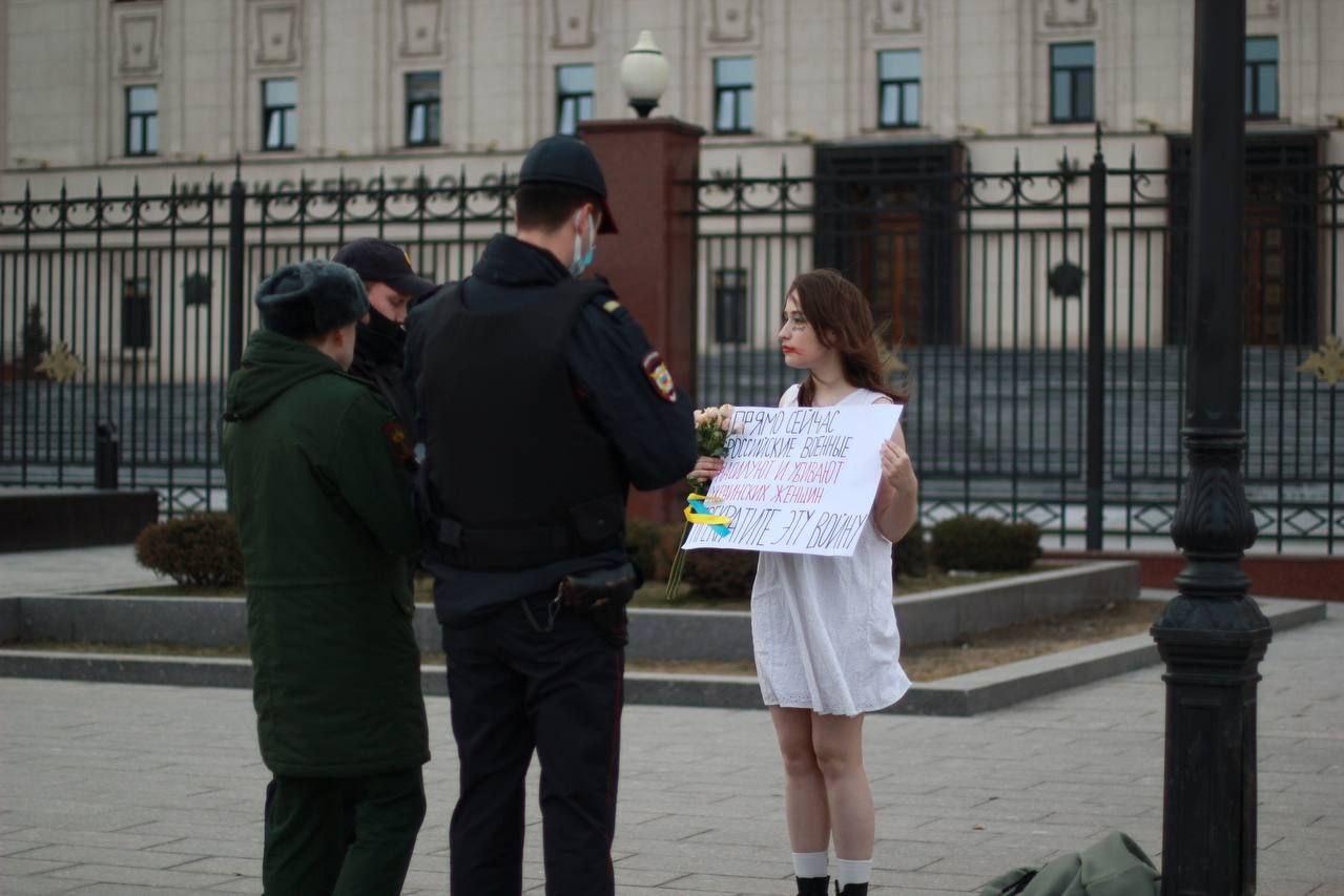 Valkoisiin pukeutunut nainen seisoo kadulla pitäen kädessään venäjänkielistä tekstiä sisältävää kylttiä.