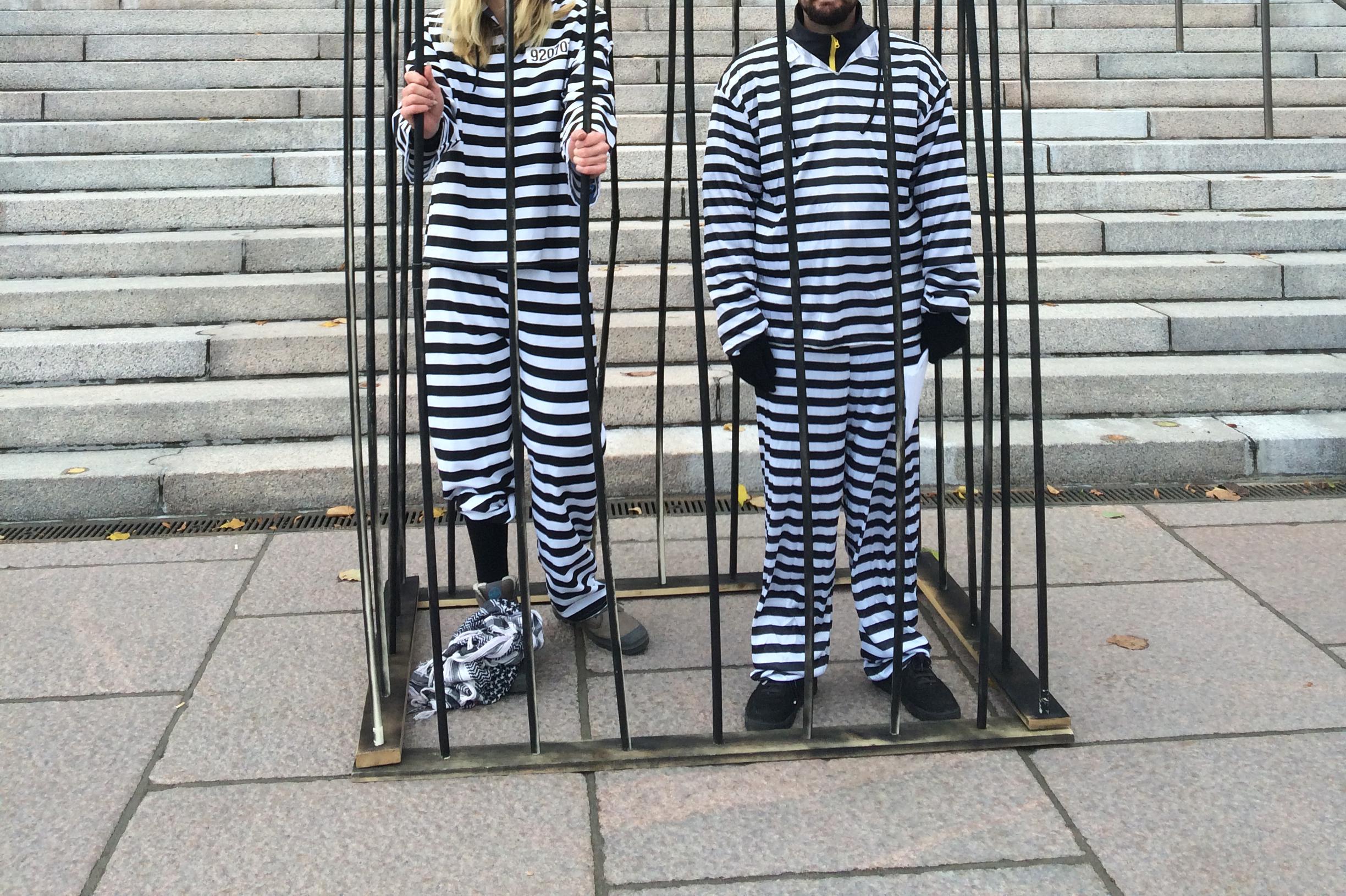 Kaksi valkomustaraidalliseen vanginasuun pukeutunutta mielenosoittajaa seisoo häkissä eduskuntatalon portaiden edessä.